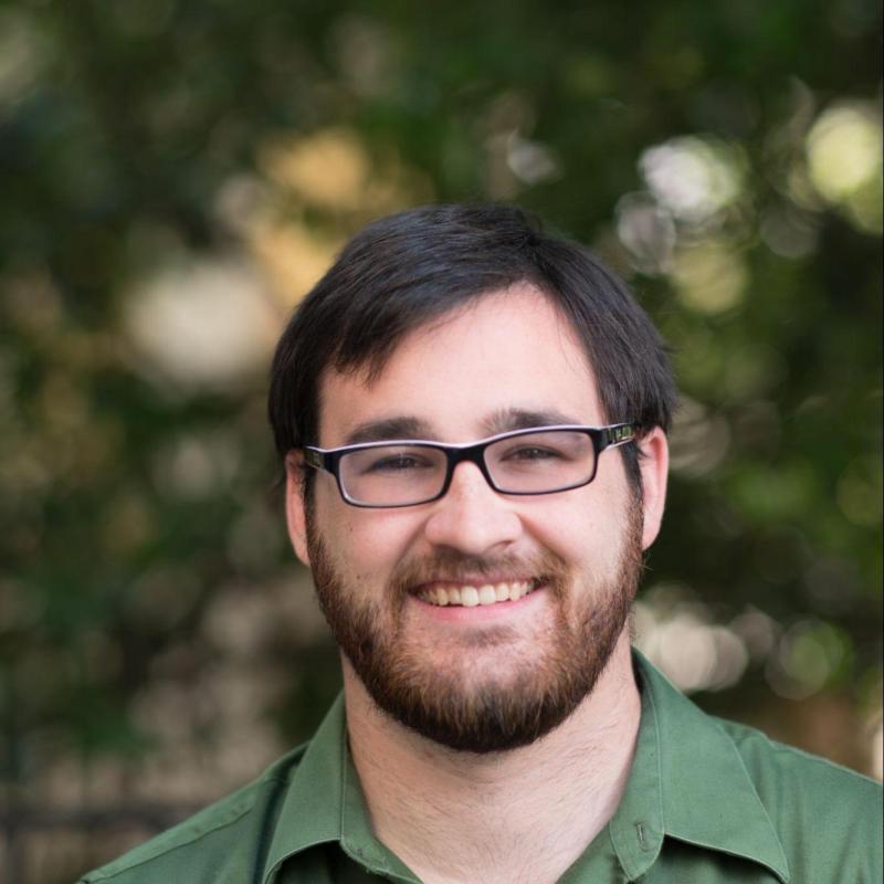 Man with glasses, beard, and green shirt smiling outdoors.