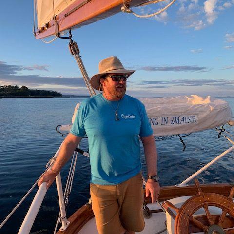 Man in a hat steering a sailboat at sunset on calm water.