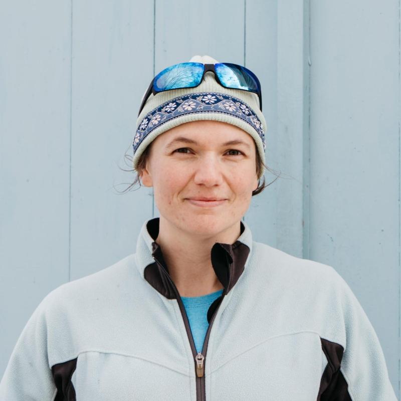 Person wearing a winter hat and sunglasses on head, standing against a light blue wall.