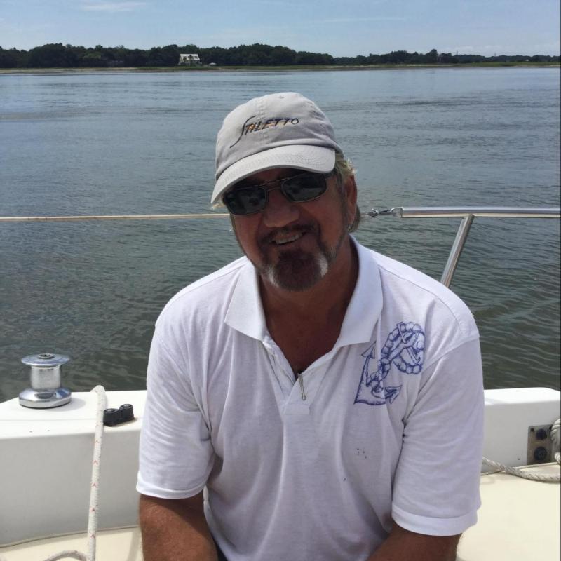 Man sitting on a boat, wearing a hat and sunglasses, with water and trees in the background.